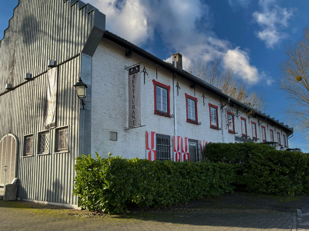 Foto von einem weißen Gebäude mit roten Fensterrahmen und Restaurantschild. Davor Büsche, links Metallwand und Kopfsteinpflaster.