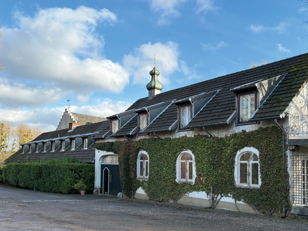 Foto von einem langen, weißen Gebäude mit Efeuwuchs, dunklem Dach, kleinen Gauben und Kuppel, daneben Büsche unter blauem Himmel.